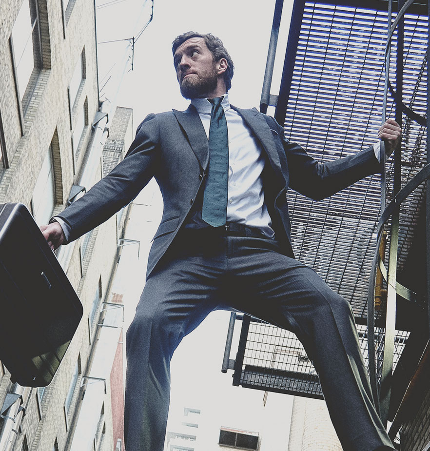 Man hanging in tactical Grayman and Company suit with briefcase hanging from fire escape